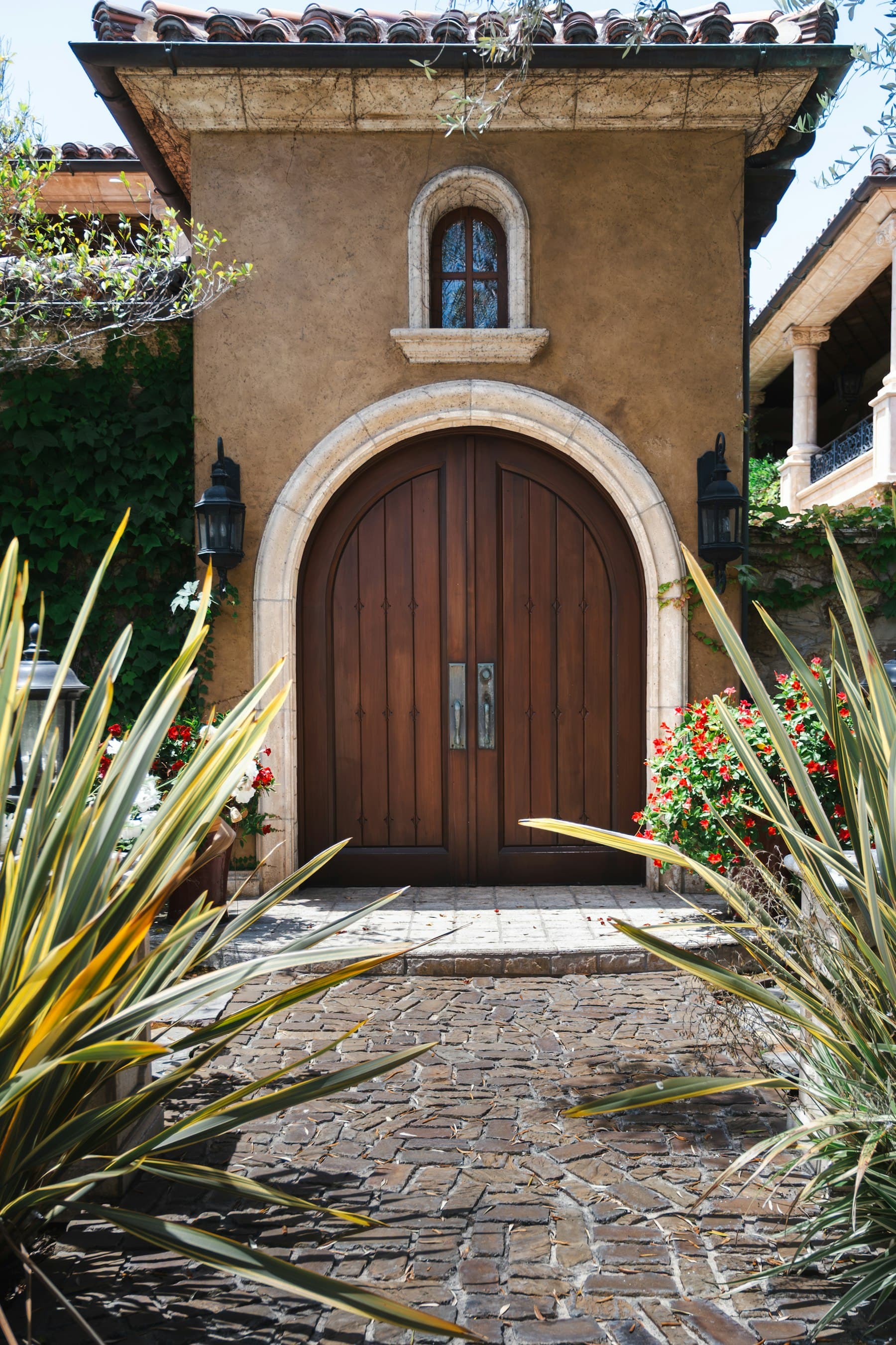 Arched wooden double doors in a carved stone surround, flanked by lanterns and lush plantings